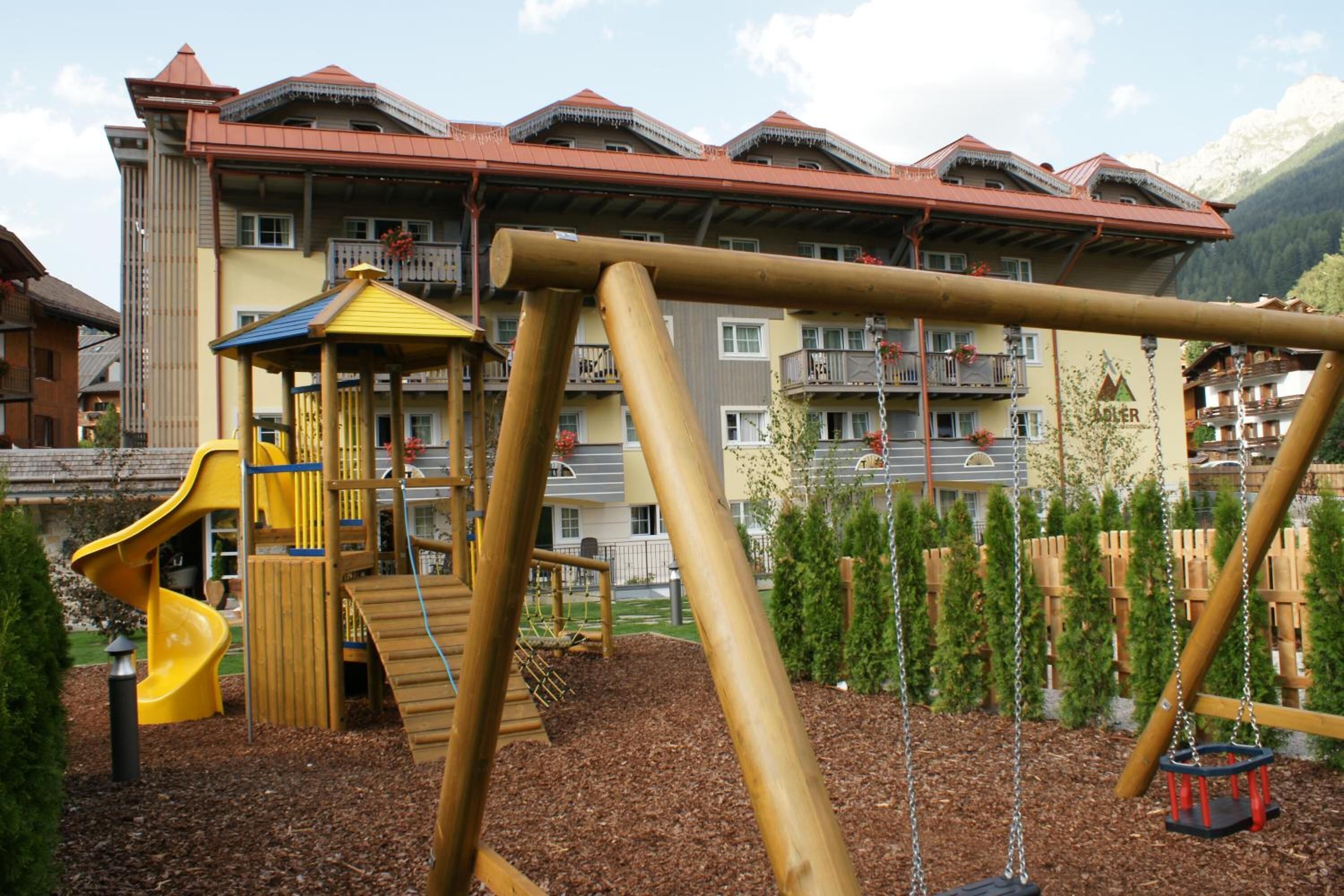 Un parque infantil con un tobogán amarillo y columpios, rodeado por una cerca de madera y un edificio con balcones y un techo rojo.