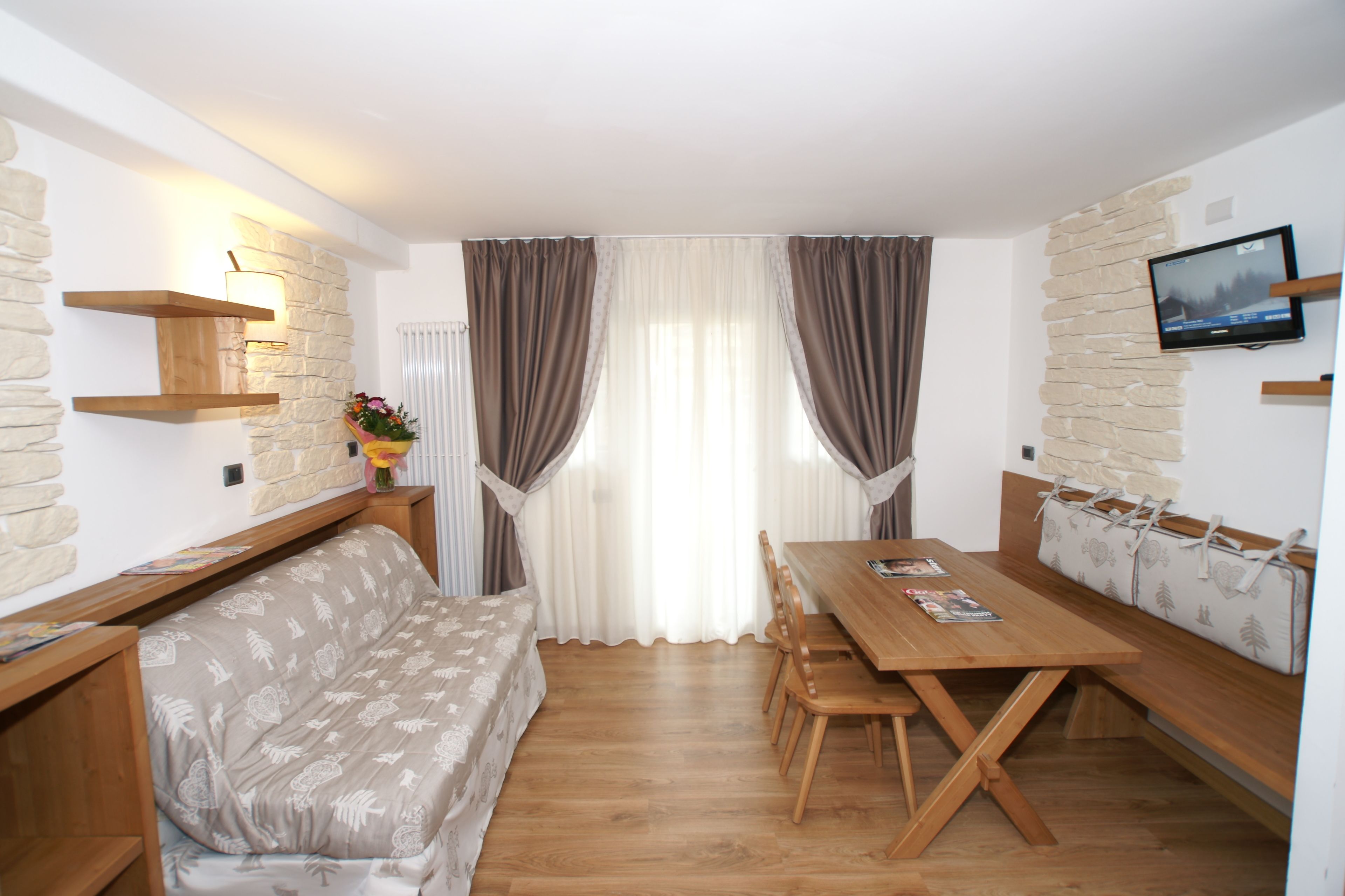 A cozy room with a sofa, wooden dining table, and benches. There is a TV mounted on the wall and a stone accent wall.