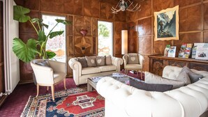 Een kamer met een grote plant, een beige bank, een witte fauteuil en een houten kast met boeken en een ingelijst beeld.