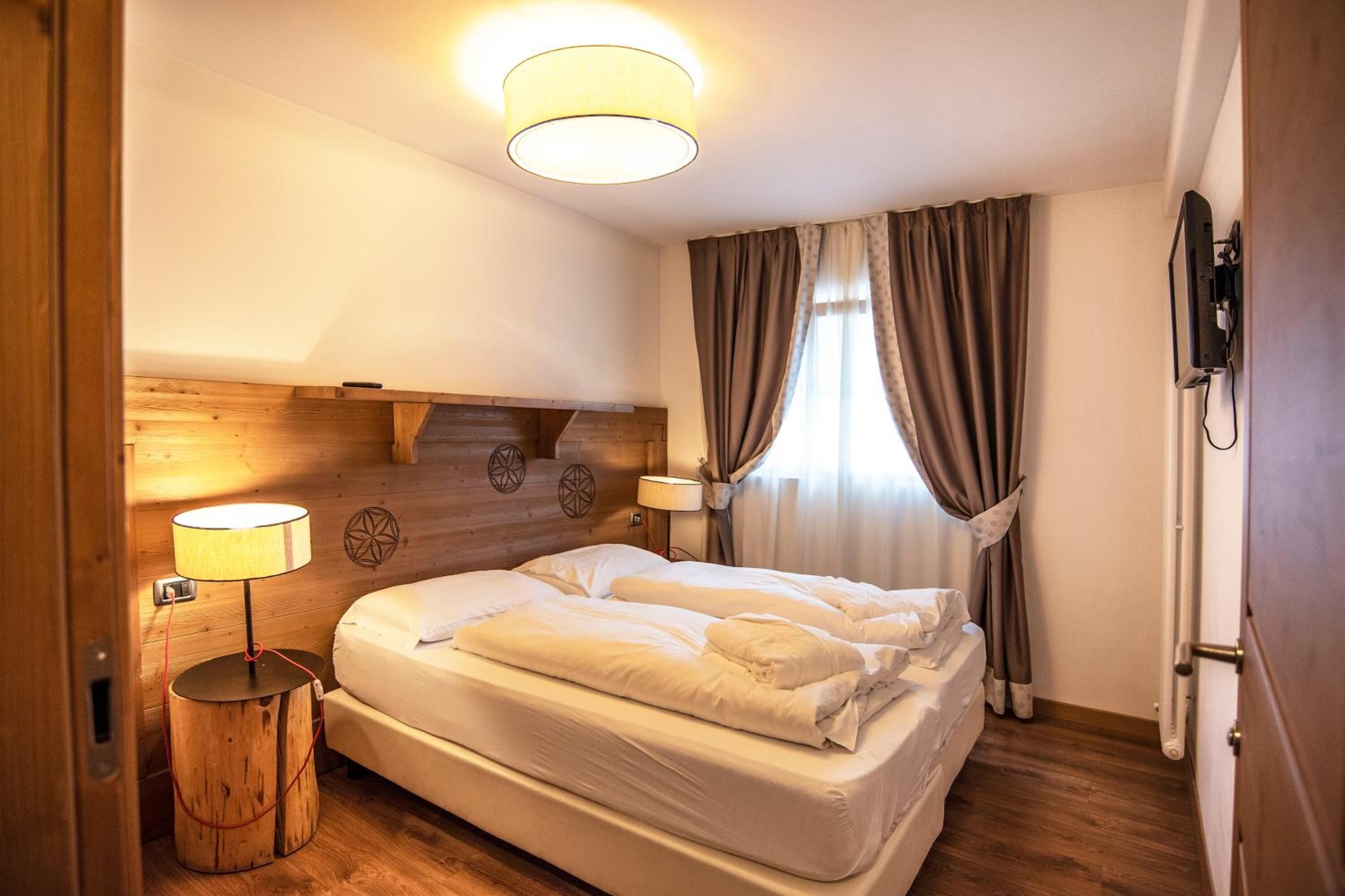 A hotel room with a wooden headboard, two beds, a round bedside table with a lamp, and a television mounted on the wall.