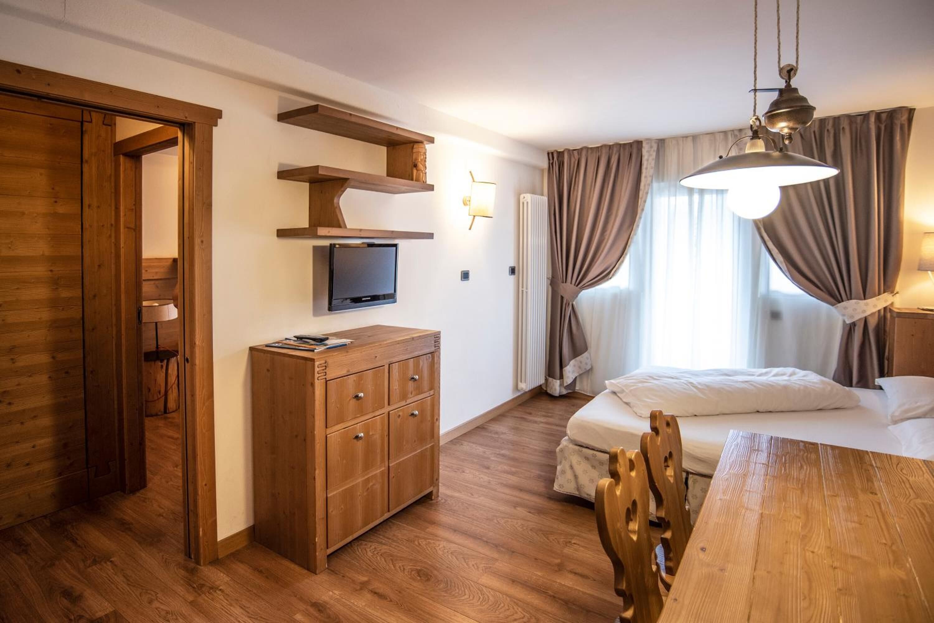 A hotel room with a wooden floor, a bed with white linens, a wooden dresser, a television on a shelf, and a hanging light fixture.