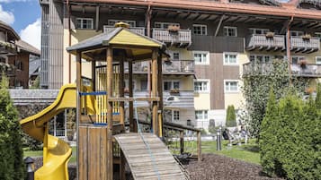 A playground with a yellow slide and a wooden climbing structure, surrounded by a landscaped garden with mulch and trees.