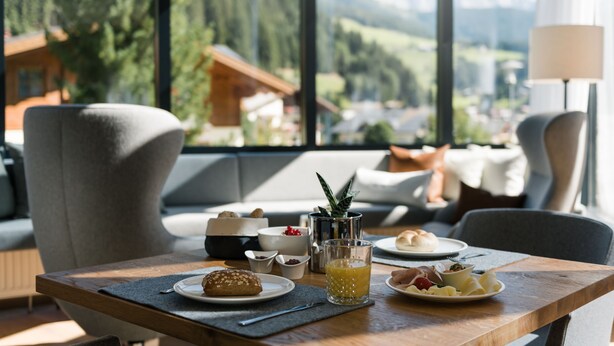 A breakfast table with plates of food, a glass of juice, and a view of mountains through large windows. - meiZeit Lodge (Filzmoos)