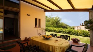 Un patio al aire libre con una mesa dispuesta para comer, rodeado de sillas y vistas a un jardín y edificios al fondo.