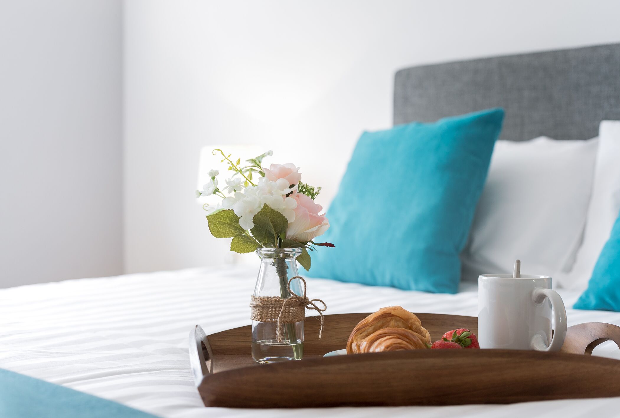 A neatly made bed with a tray containing a croissant, a strawberry, and a white mug. A small vase with flowers is placed on the bed.