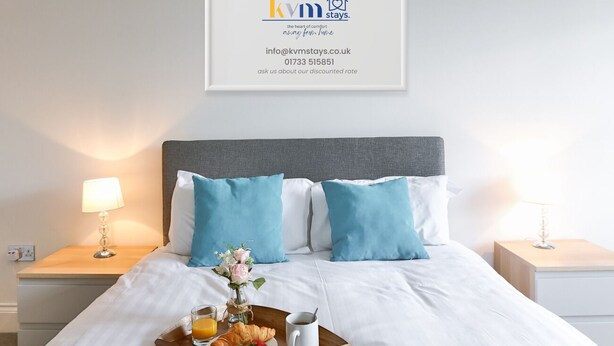 A neatly made bed with a tray of breakfast, a framed logo above, and a bedside lamp on each side. - KVM - Jubilee Mansions 2 (Peterborough)