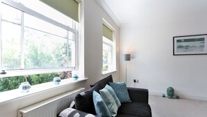 A modern living room with a dark grey sofa, two windows with white frames, a radiator, and a framed picture on the wall. - KVM - Jubilee Mansions 2 (Peterborough)