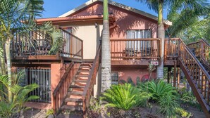 A two-story building with a balcony on the upper floor, a staircase leading to the upper level, and a small garden area with tropical plants. - Elephant Coast Guest House (St. Lucia)