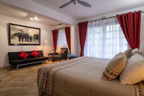 A bedroom with a large bed, a sofa, a chair, and a desk. There is a ceiling fan, a window with curtains, and a wall-mounted air conditioner. - Elephant Coast Guest House (St. Lucia)