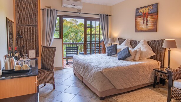 A bedroom with a large bed, a desk with a coffee maker, a chair, and a balcony with a view of trees. - Elephant Coast Guest House (St. Lucia)