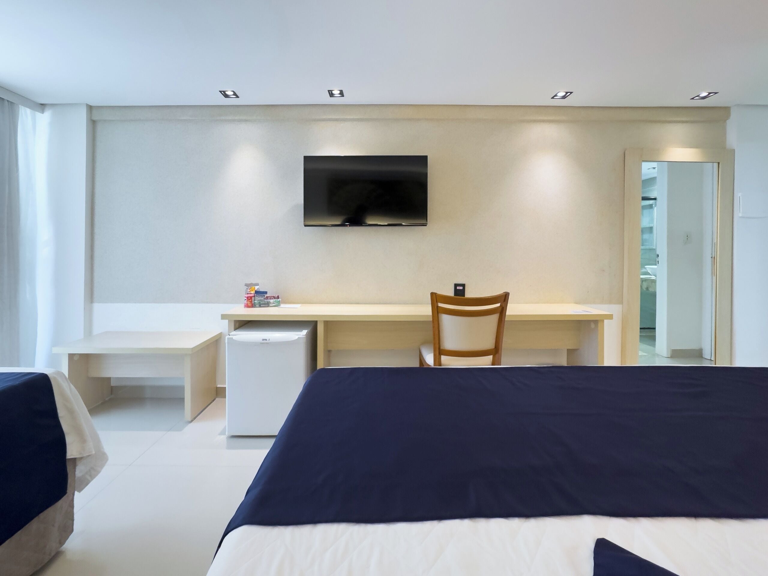 Una habitación de hotel moderna con una cama, un escritorio, una silla, un refrigerador pequeño y un televisor montado en la pared.