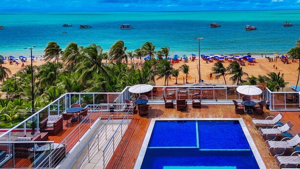 Zona junto a la piscina con sillas de descanso y sombrillas, con vista a una playa con palmeras y barcos.