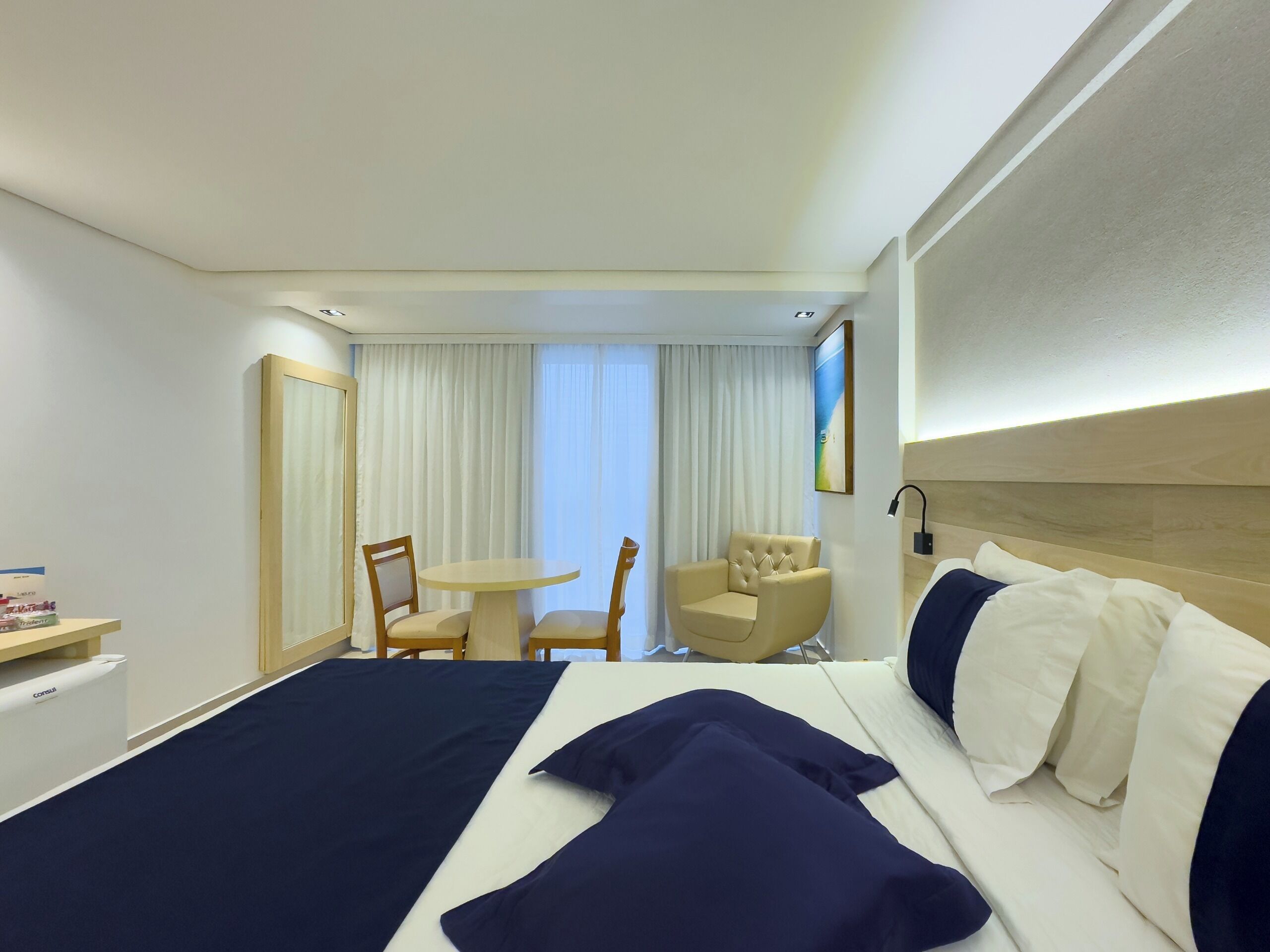 A hotel room with a large bed, a bedside table, a chair, a small table, and a window with curtains.