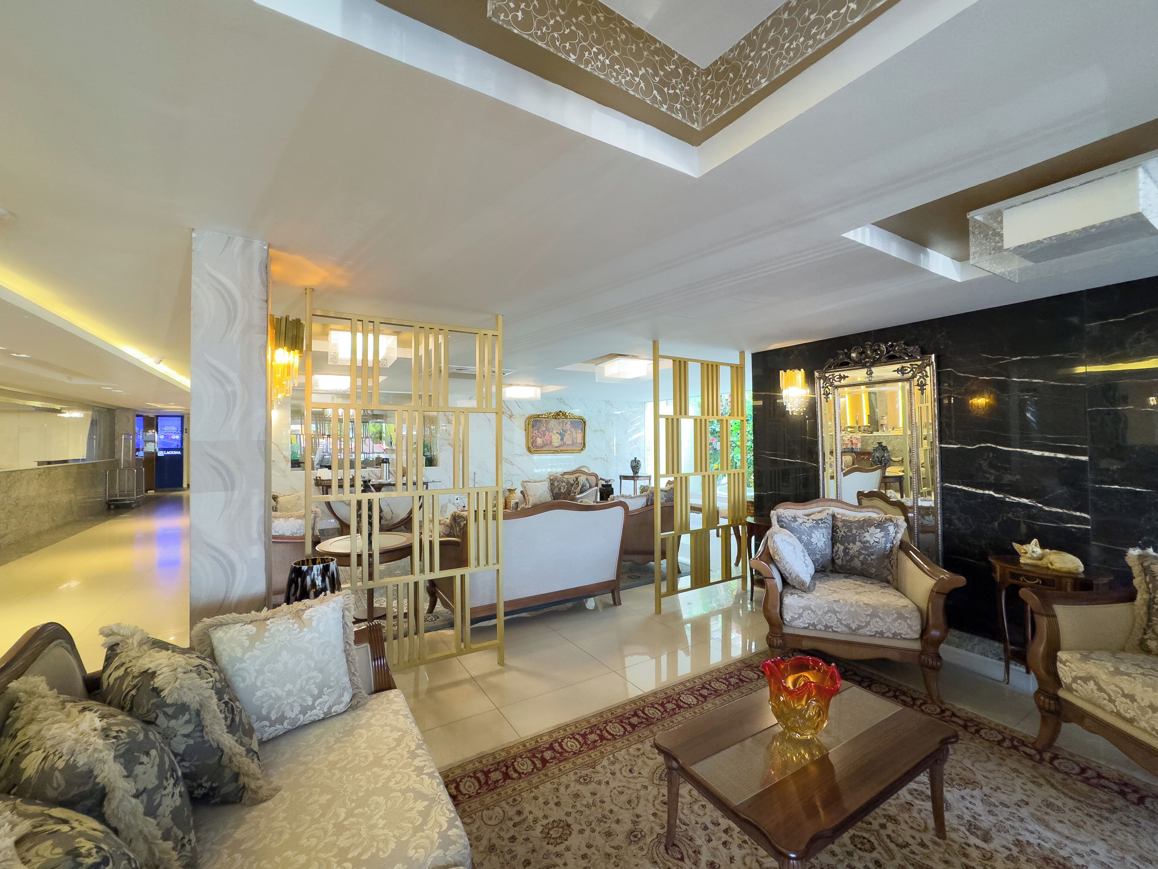 A hotel lobby with a reception desk, seating area with sofas and armchairs, a decorative mirror, and a patterned rug.