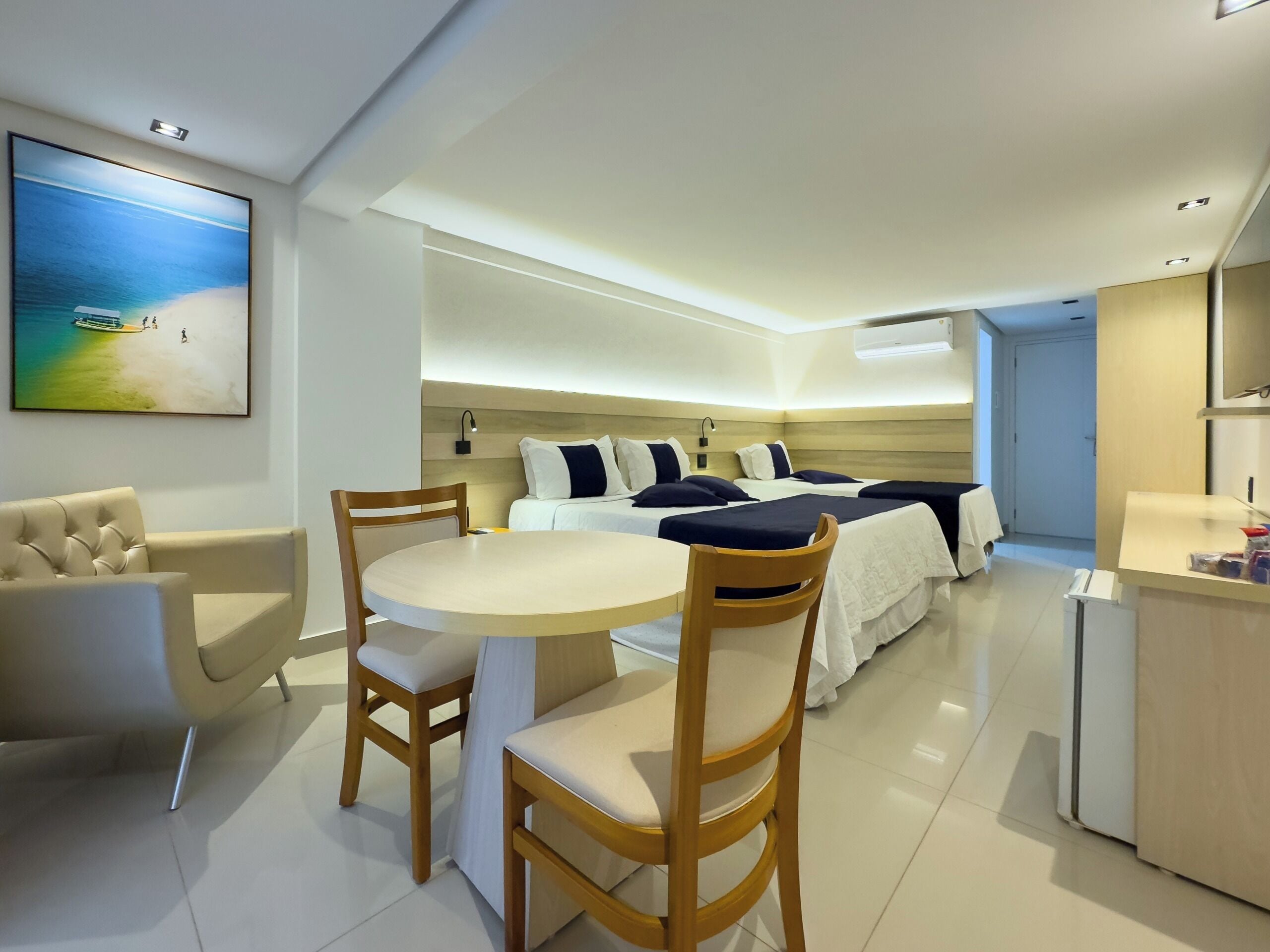 A modern hotel room with a dining area, a bed with multiple pillows, a wooden headboard, and a painting of a beach scene.