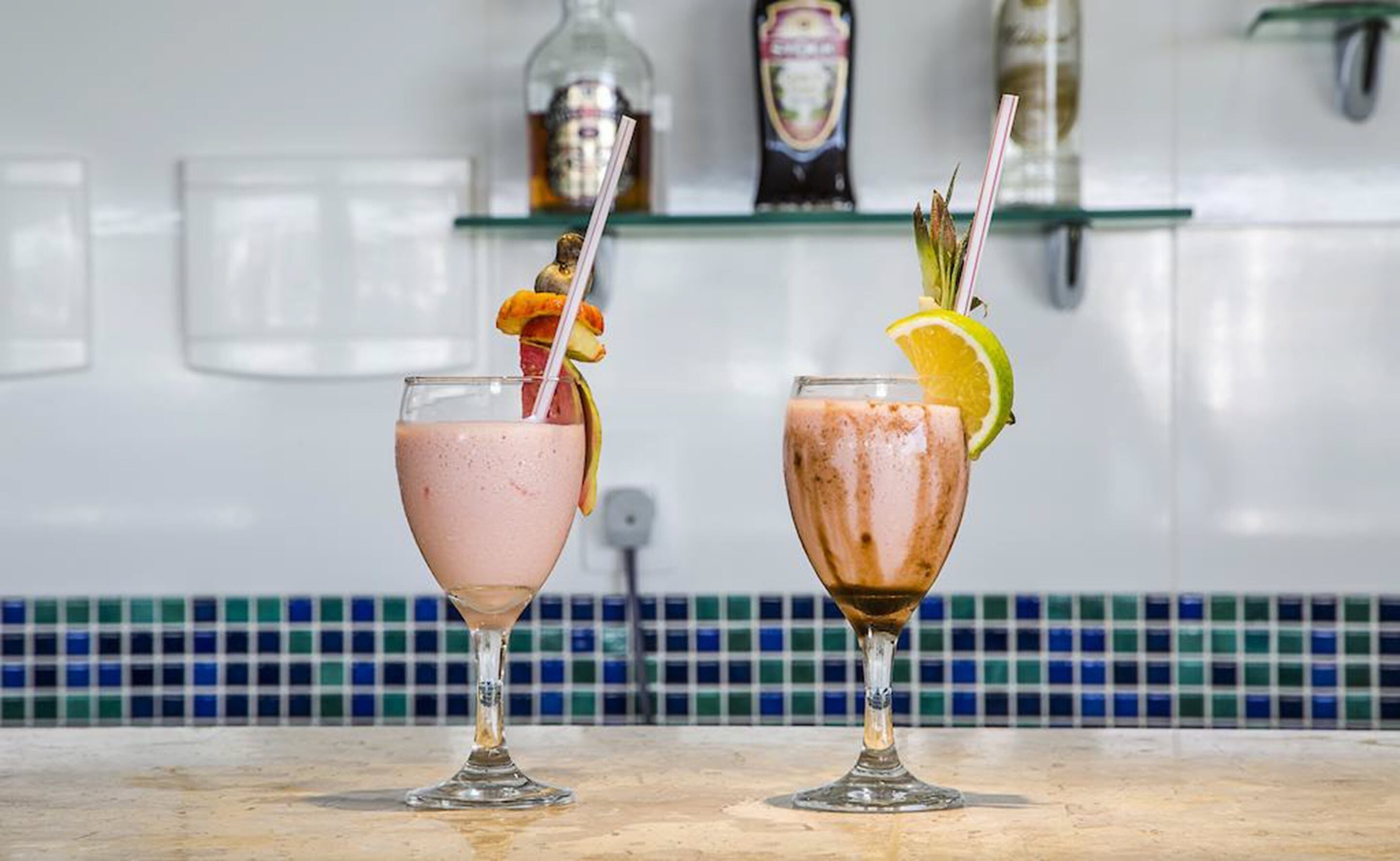 Two drinks in stemmed glasses with fruit garnishes on a kitchen counter.