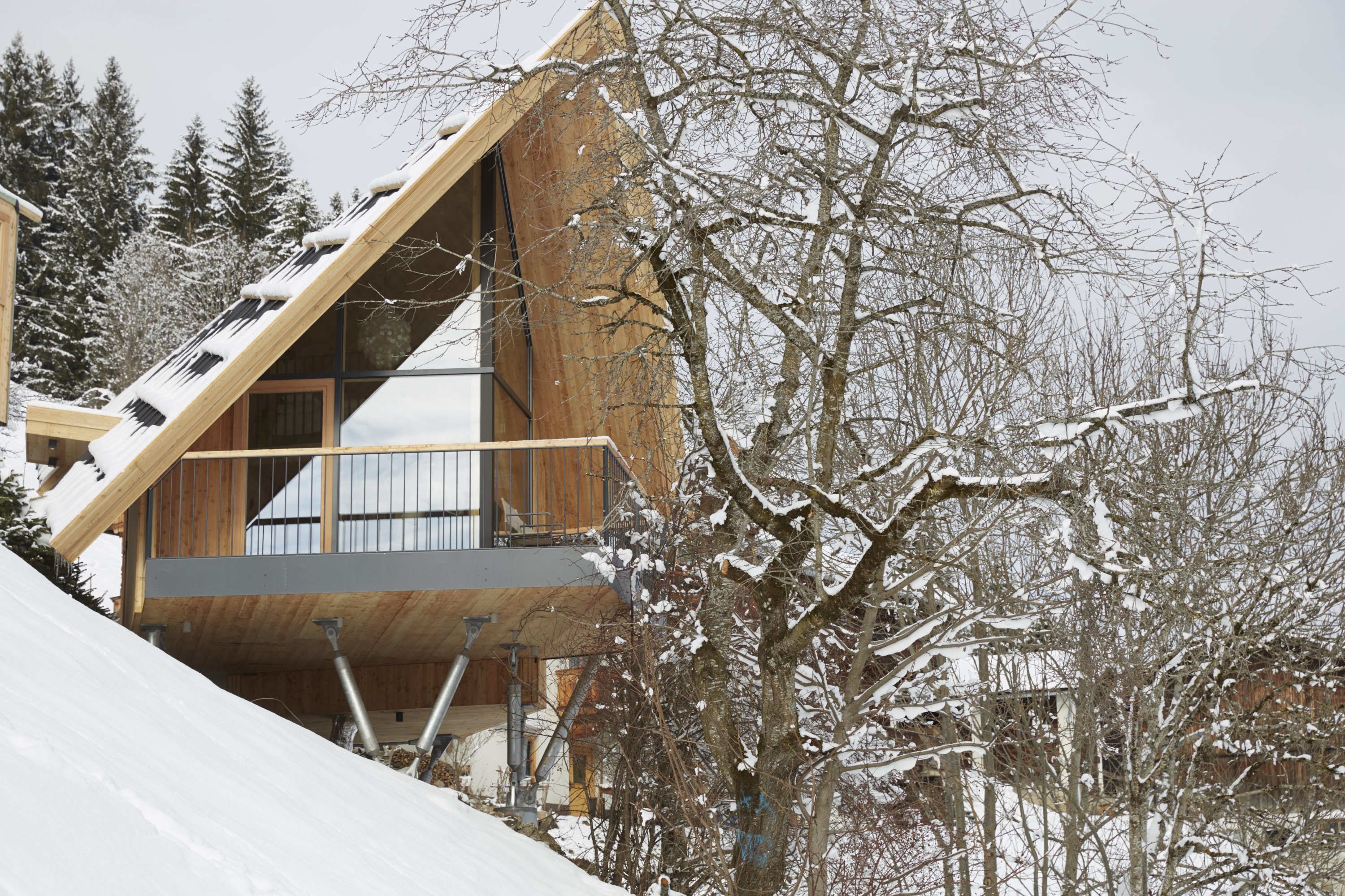 A modern wooden cabin with a sloped roof, a balcony, and a view of snow-covered trees.