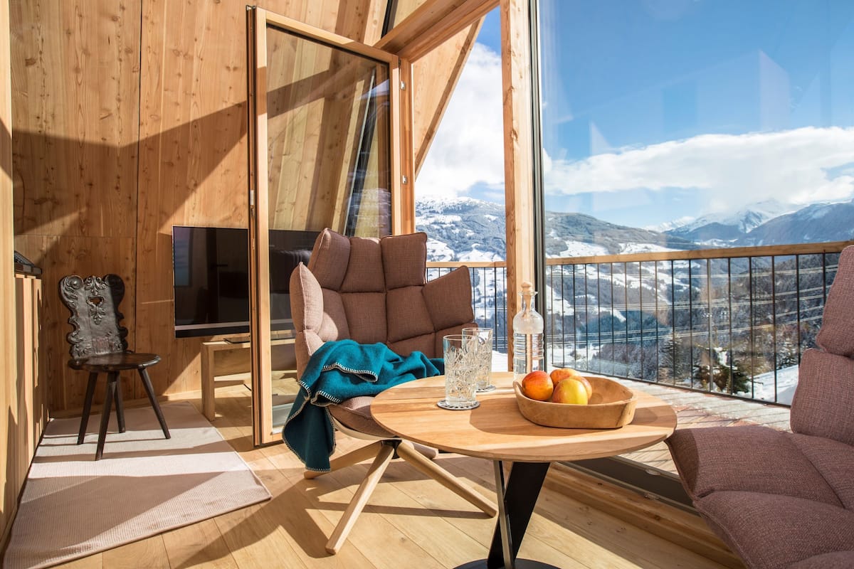 A wooden interior with a balcony, a television, a chair, a table with fruit, and a view of snow-capped mountains.