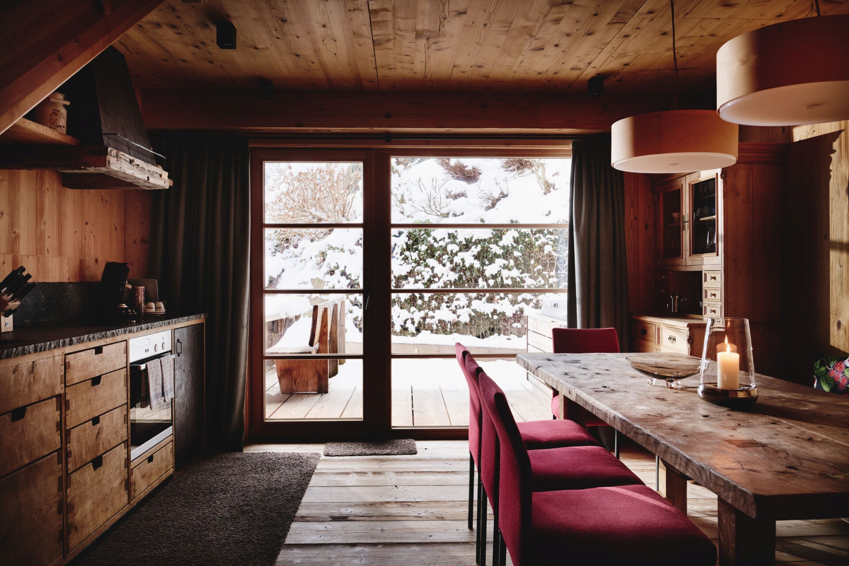 A wooden cabin interior with a large window, a wooden dining table, and red chairs.