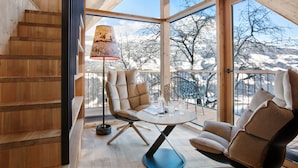 A wooden interior with a staircase, a round table with chairs, and a view of snow-covered mountains through large windows.