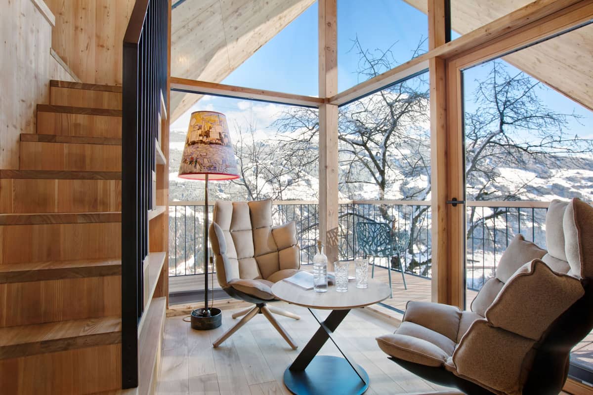 A wooden interior with a staircase, a round table with chairs, and a view of snow-covered mountains through large windows.