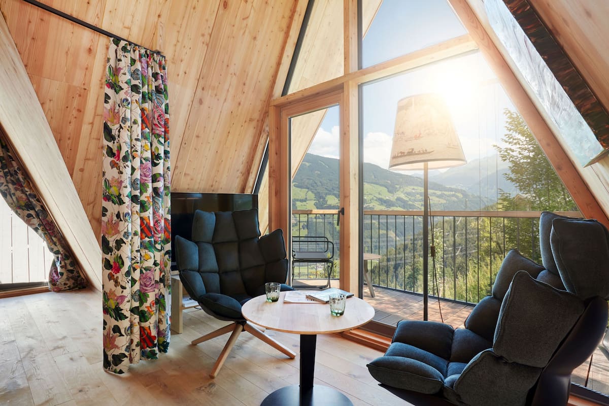 A cozy room with a wooden ceiling, a round table, two chairs, and a balcony with a view of the countryside.