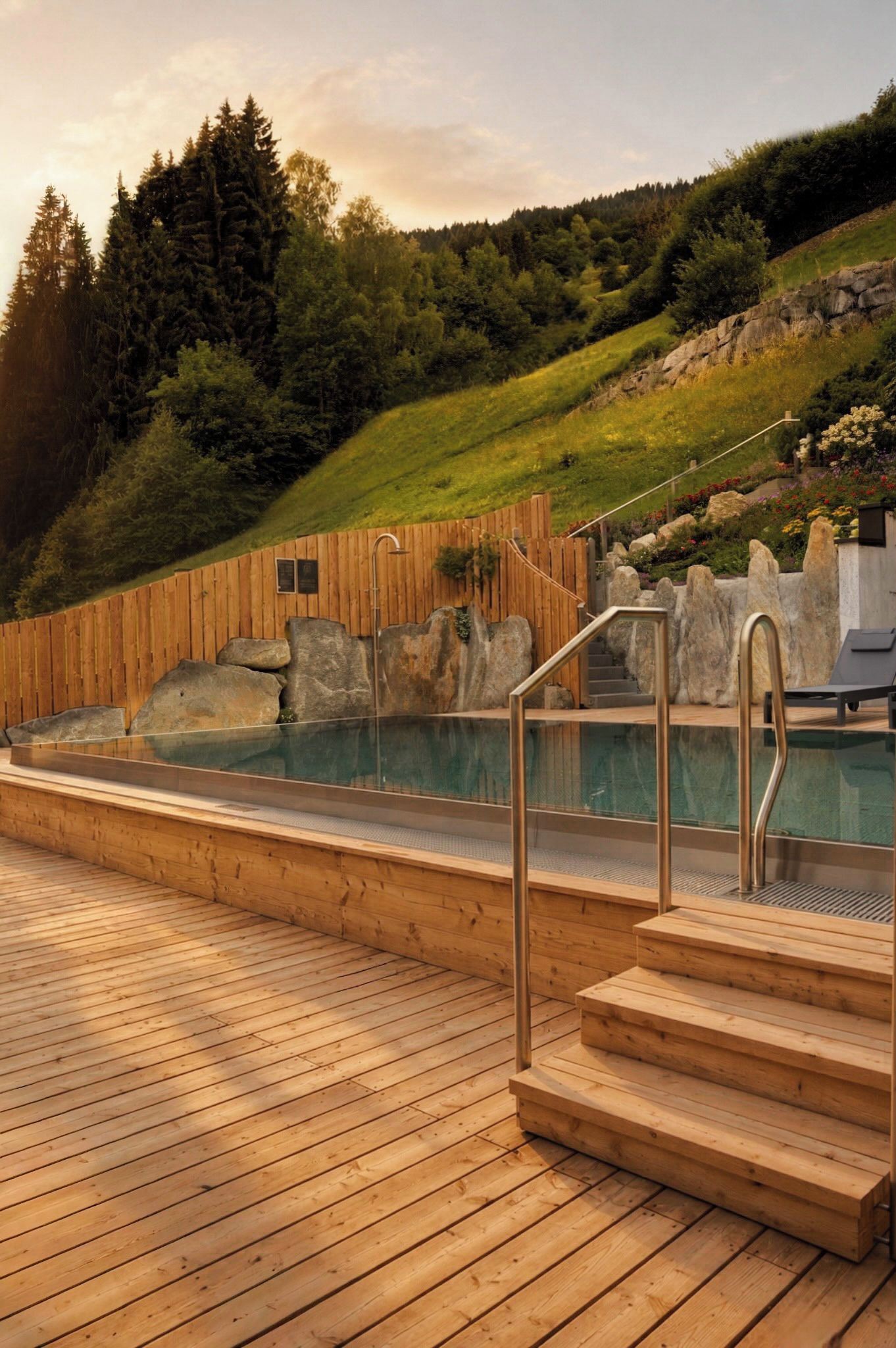 Eine Holzterrasse führt zu einem Schwimmbecken, im Hintergrund eine Steinmauer und ein Holzzaun.