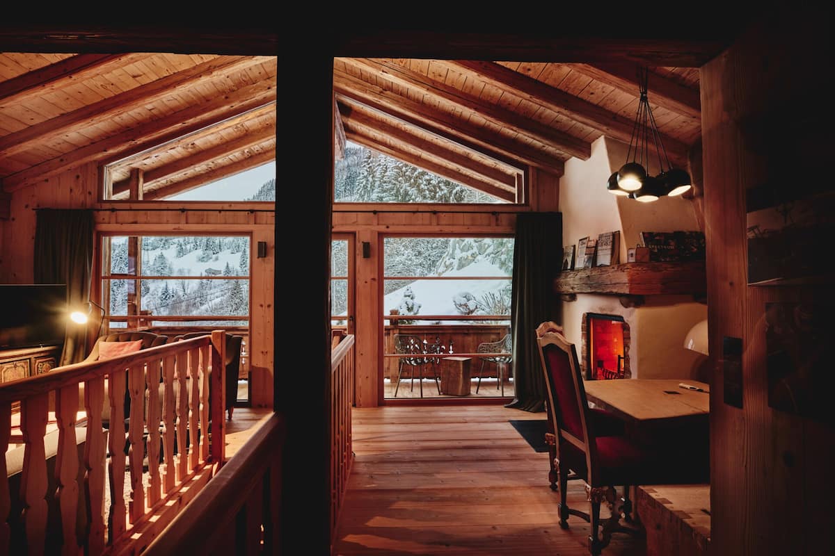A cozy cabin interior with a wooden interior, a dining area with a table and chairs, and a view of snow-covered trees through large windows.