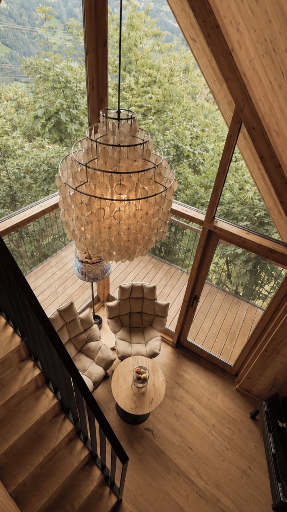 A modern interior with a wooden staircase, a round table with a vase, and a large window overlooking a forested area.