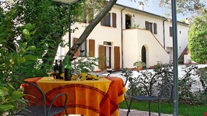 Un área de comedor al aire libre con una mesa dispuesta para dos personas, rodeada de follaje y un edificio al fondo.