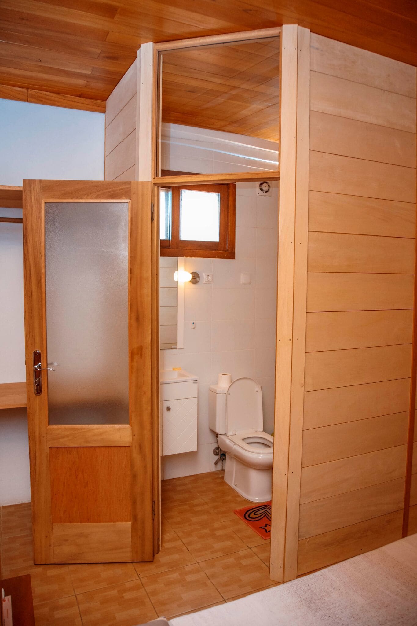 Un bagno piccolo e accogliente, con arredi in legno, porta in vetro satinato, un water bianco e una piccola finestra sopra il lavabo.