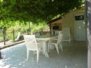 Une terrasse extérieure avec une table ronde et des chaises blanches, une cheminée en briques et une porte avec une fenêtre.