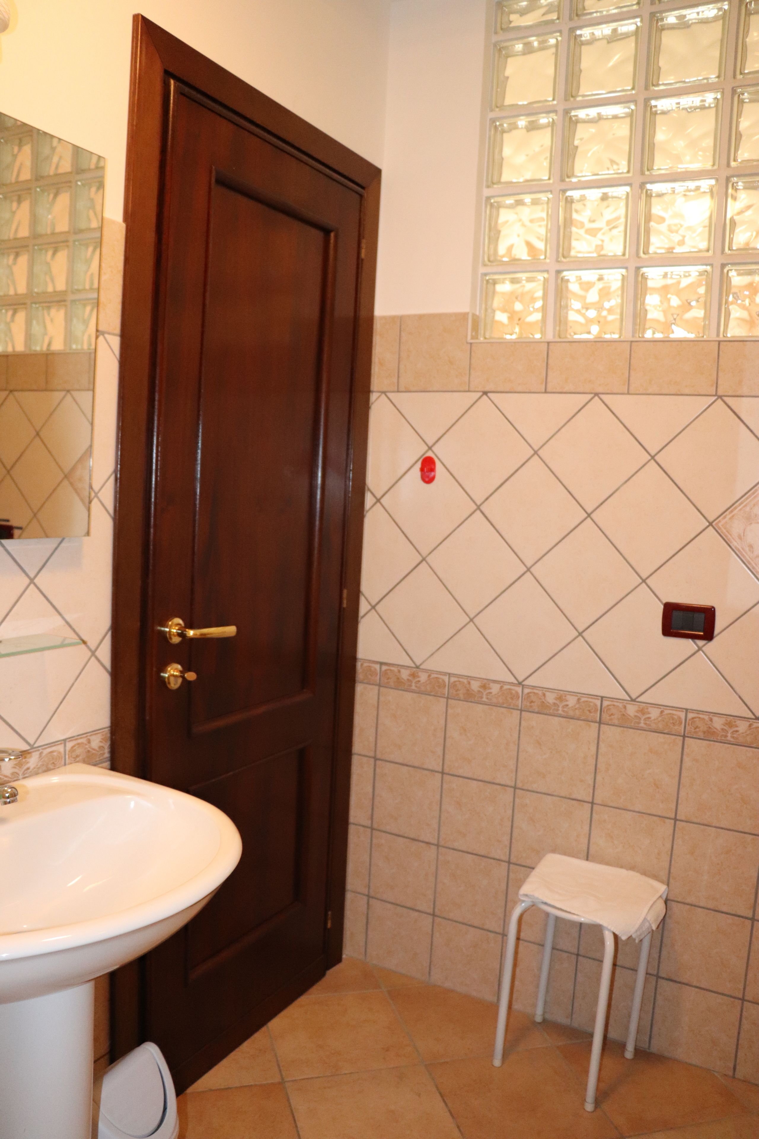 Un baño con puerta de madera, un lavamanos blanco, un taburete pequeño y una pared con azulejos y una ventana.