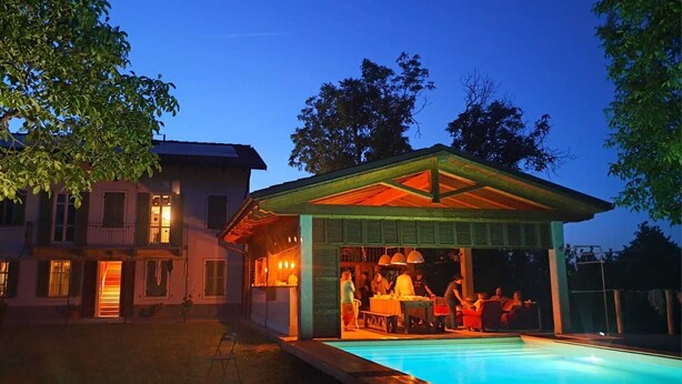 Un espace aménagé au bord de la piscine, avec un coin salon couvert et un bâtiment dont les fenêtres sont ouvertes.