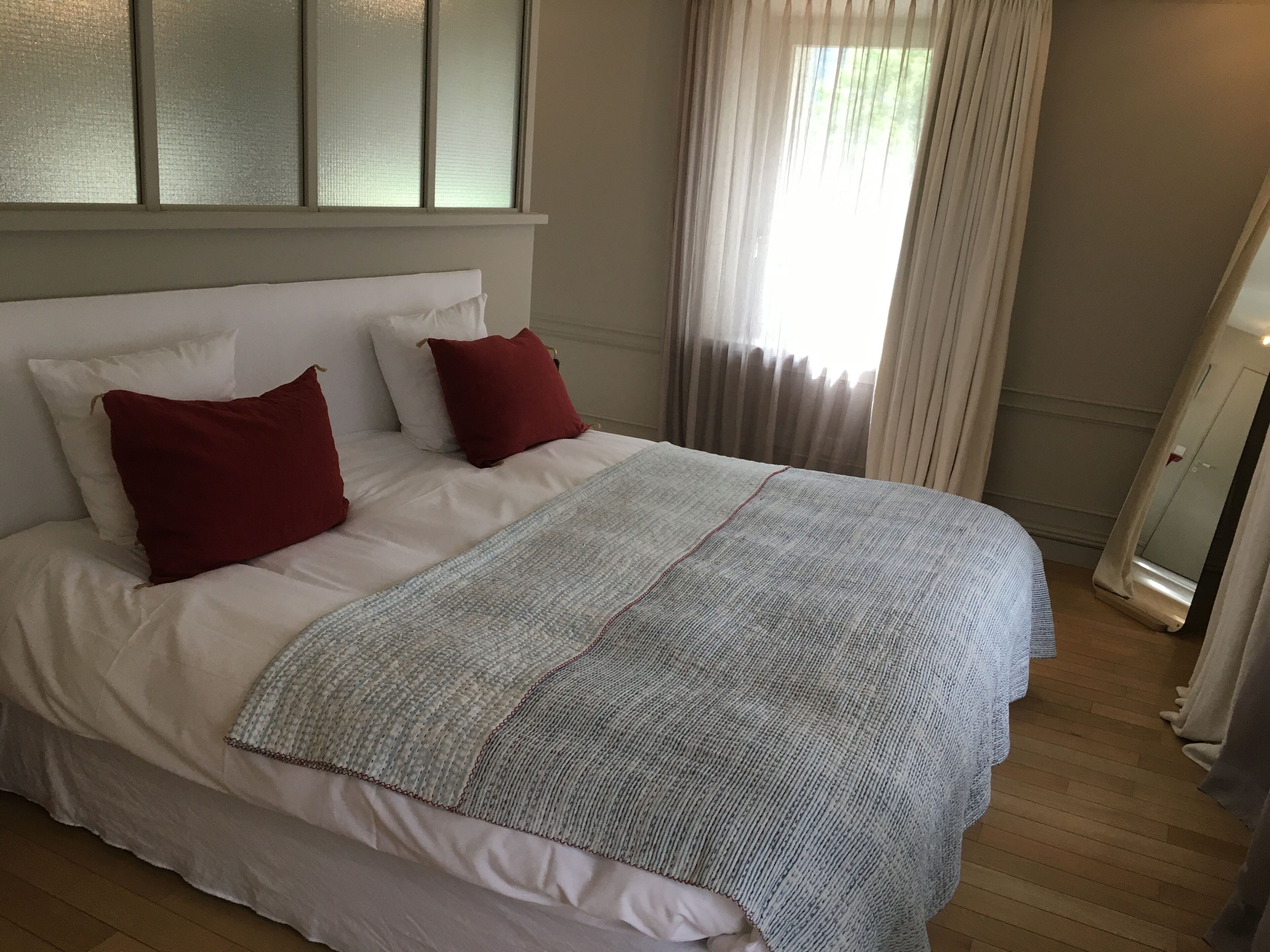 Una cama bien hecha con almohadas blancas y rojas, una manta tejida de color gris y suelo de madera.