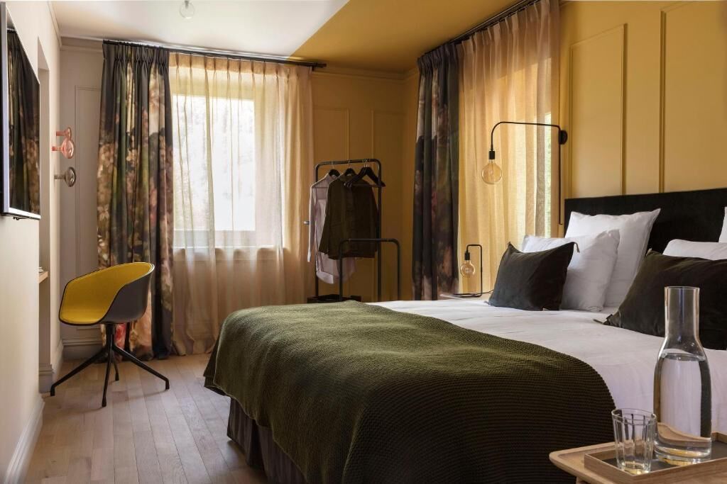 Habitación de hotel con cama, una silla, mesita de noche con lámpara y ventana con cortinas.