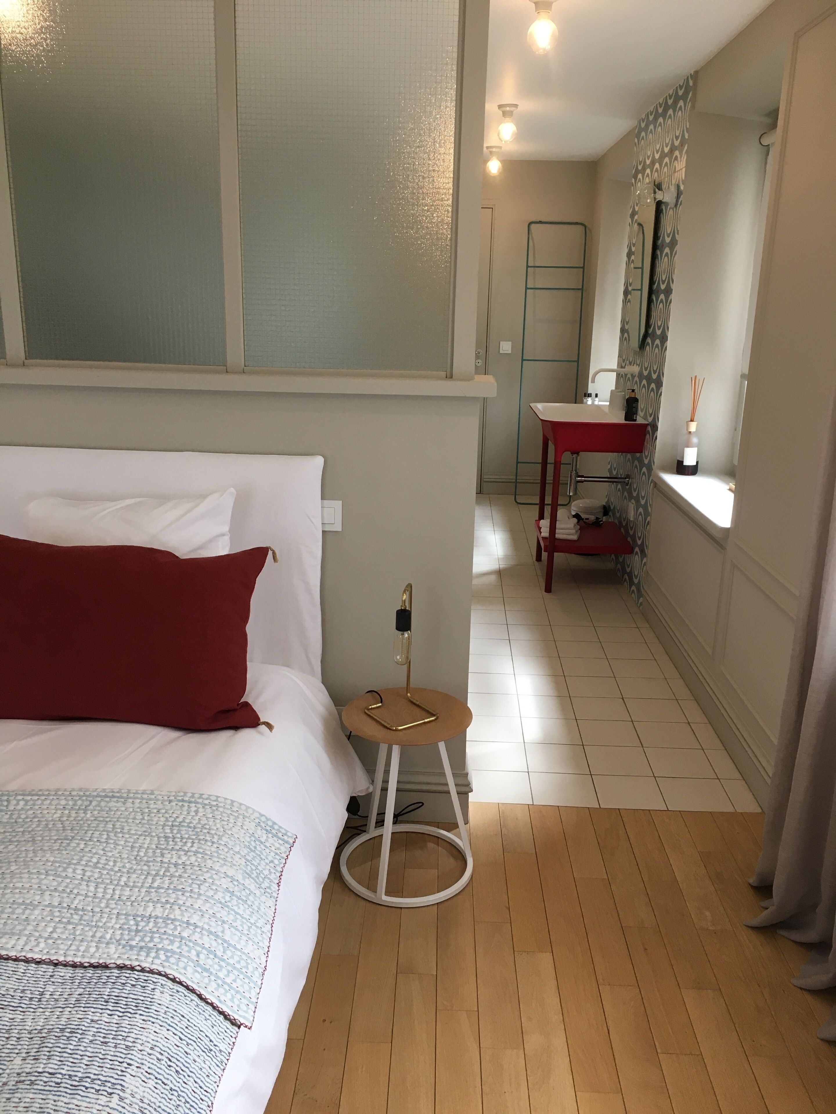Un dormitorio con cama, un taburete pequeño, una mesita de noche, una ventana con cortinas y un baño al fondo.