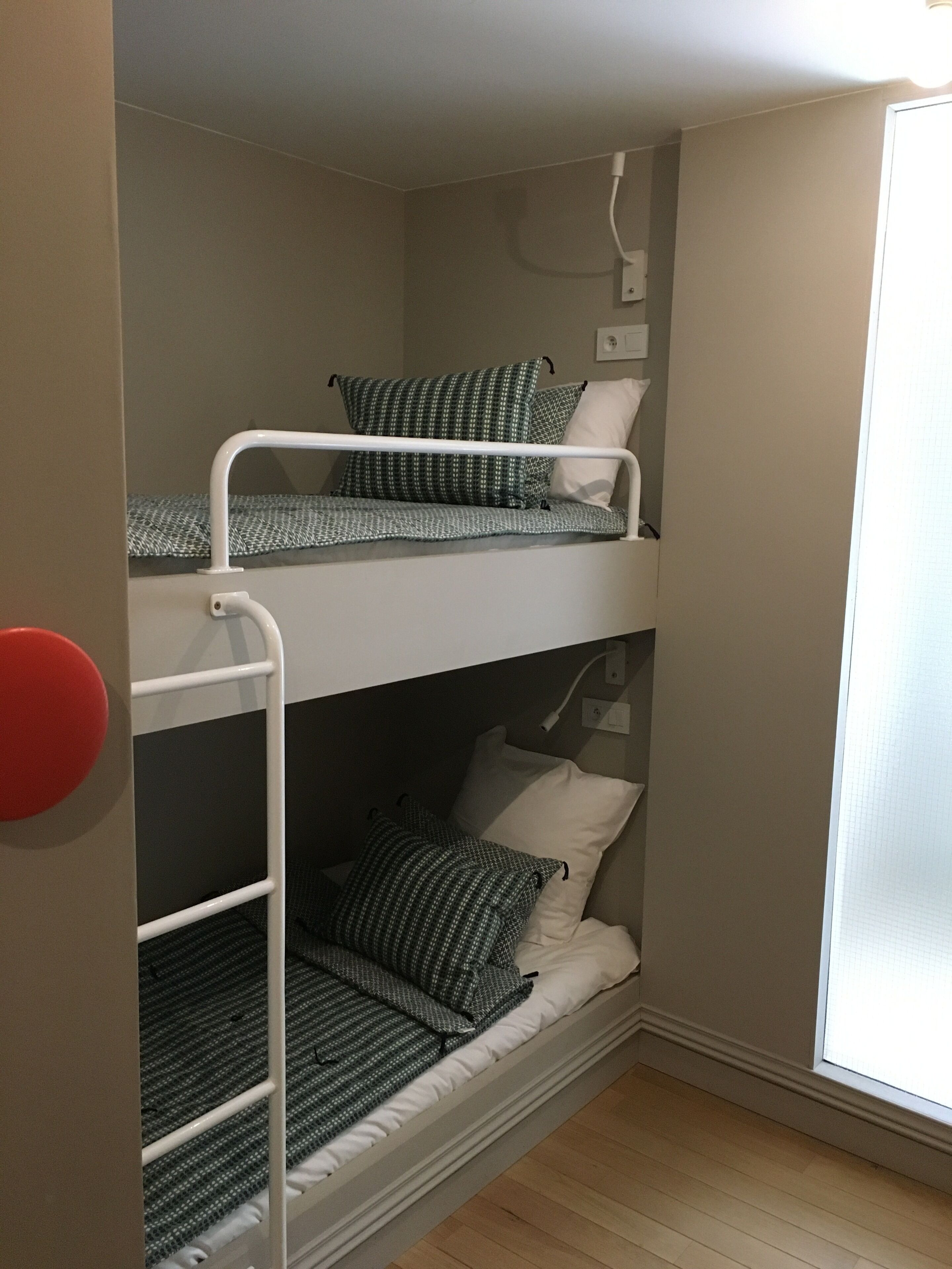 A bunk bed with two levels, each with a mattress and pillows. There is a white wall socket above the top bunk.