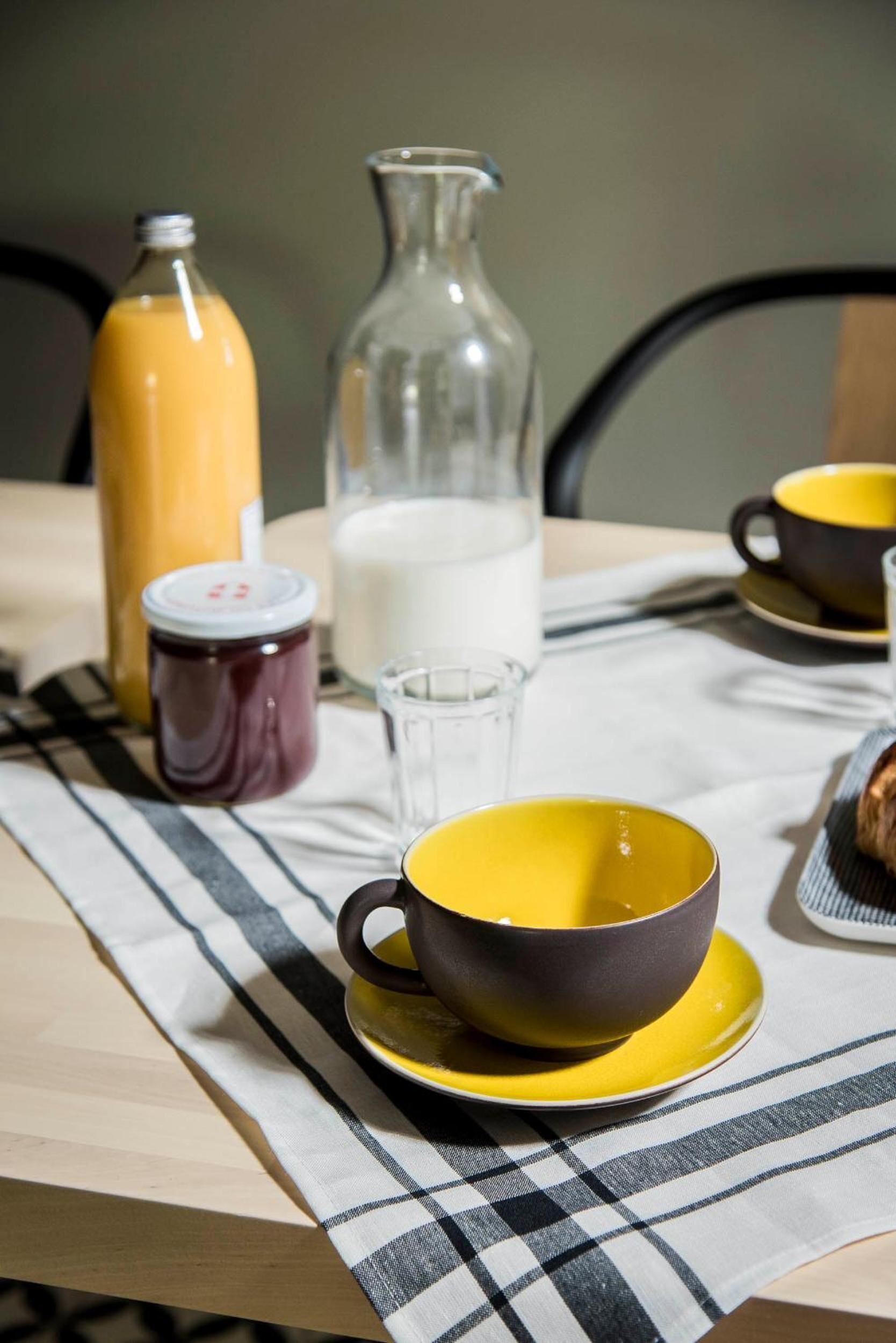 Une table à manger recouverte d’une nappe à carreaux, un bol jaune, un pichet en verre, une bouteille de jus d’orange et un pot de confiture.