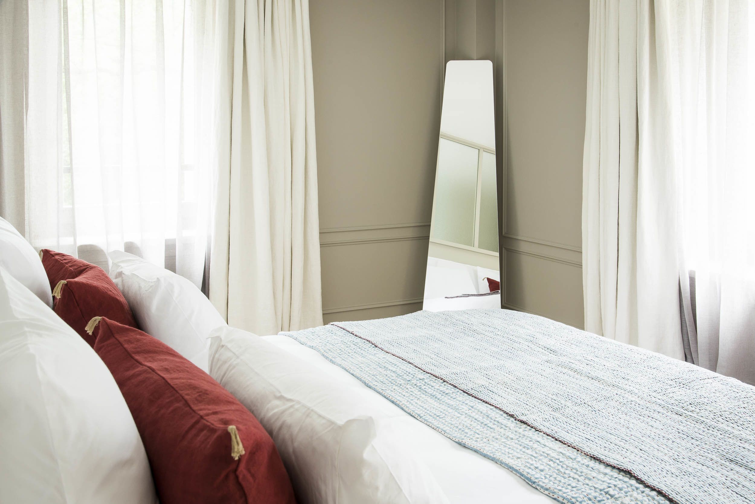 A neatly made bed with white and red pillows, a light blue blanket, and a large mirror on the wall.