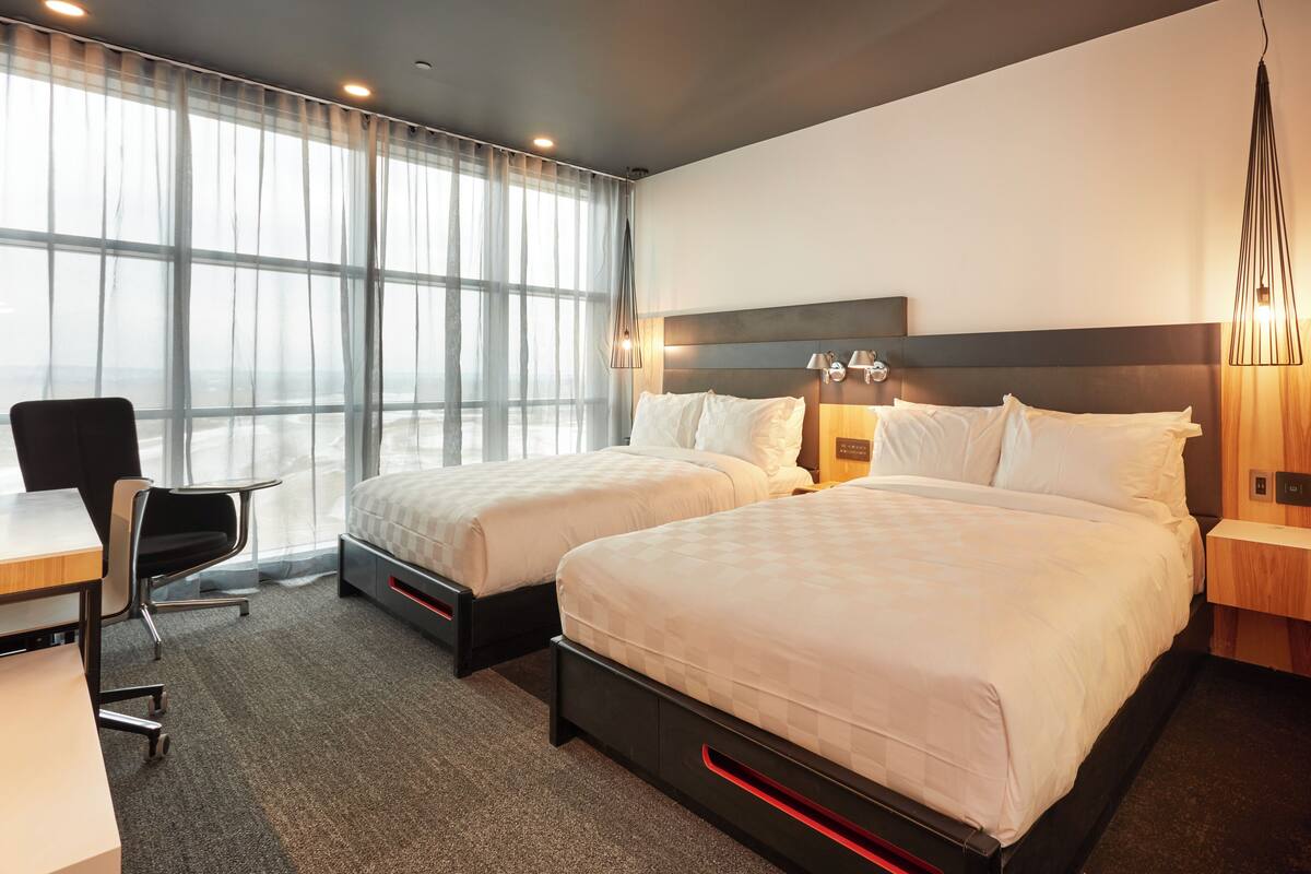 A hotel room with two beds, a desk, a chair, and a large window with sheer curtains.