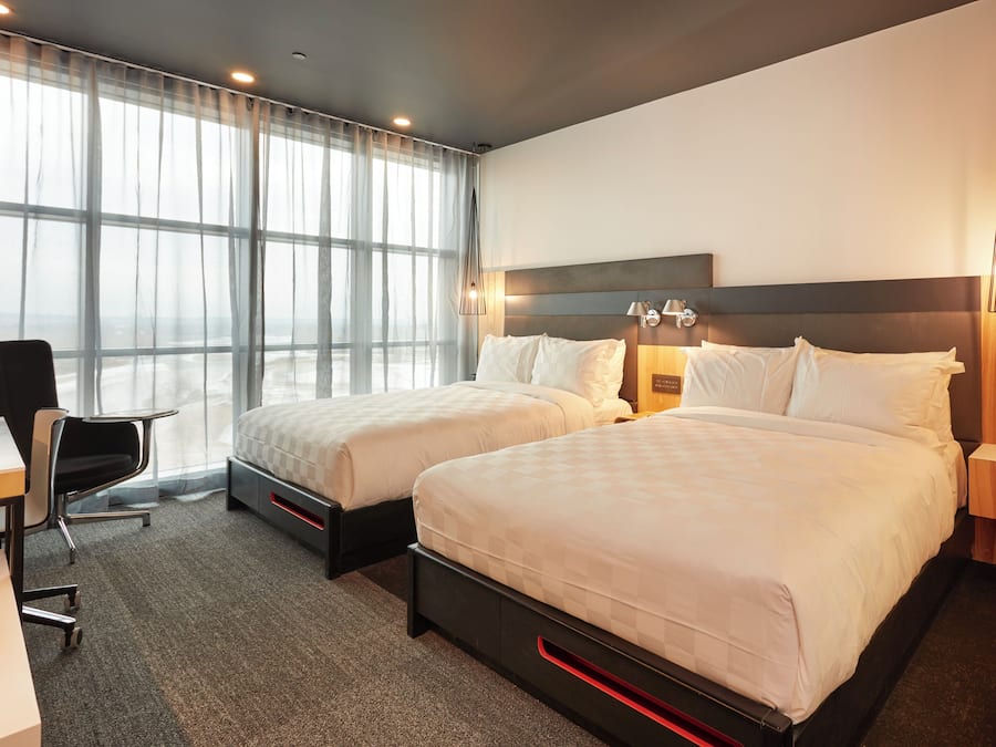 Quarto de hotel com duas camas, uma escrivaninha, uma cadeira e uma janela grande com cortinas leves.