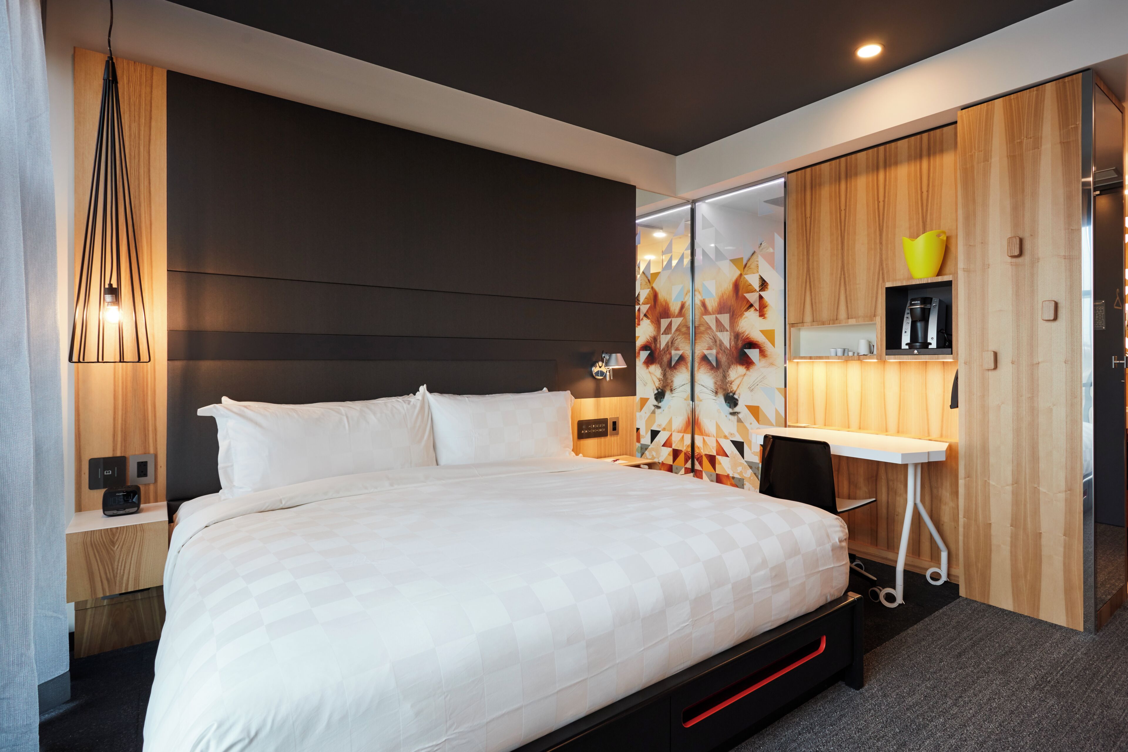 A modern hotel room with a large bed, a desk, a coffee machine, and a mirror.