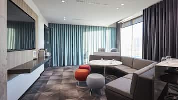 Quarto de hotel moderno com sofá, pufes, uma mesa redonda e uma grande janela com cortinas.