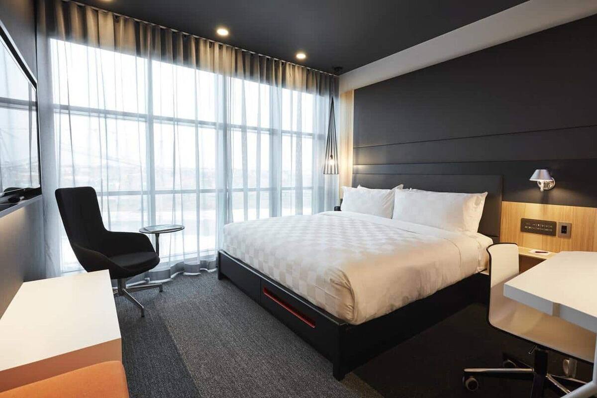 A modern hotel room with a large bed, a desk, a chair, and a window with sheer curtains.
