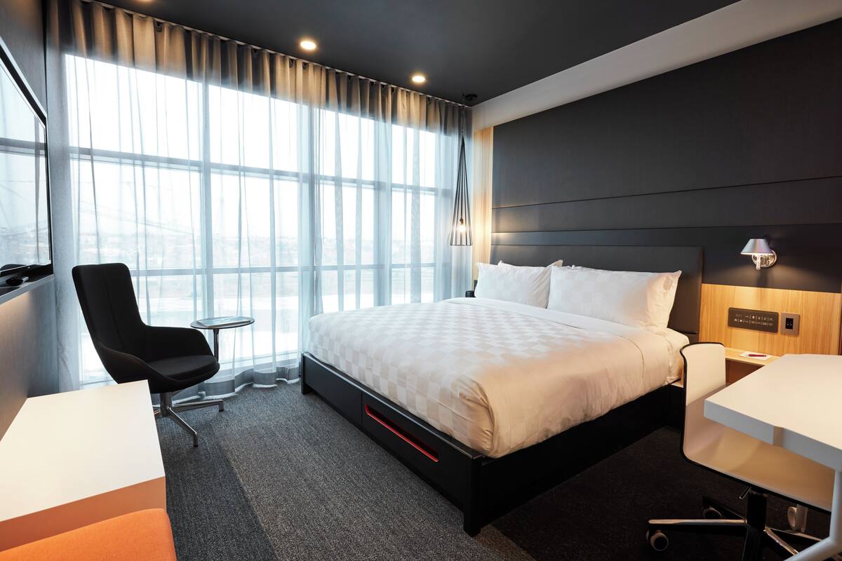 A modern hotel room with a large bed, a desk, a chair, and a view of the city through large windows.