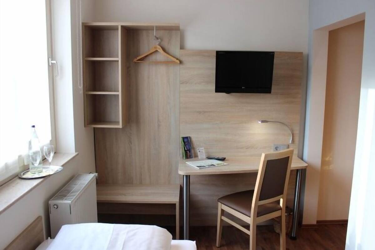 A hotel room with a bed, a desk with a chair, a TV mounted on the wall, and a wooden wardrobe with a hanging rack.