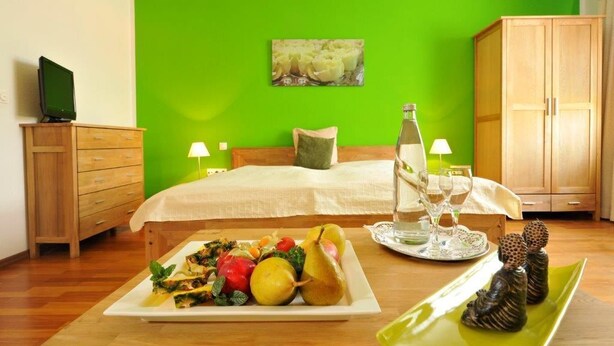 Quarto com parede verde, cama com colcha branca, cômoda de madeira, televisão sobre a cômoda, mesa de cabeceira com garrafa e copos, e um prato de fruta no chão.