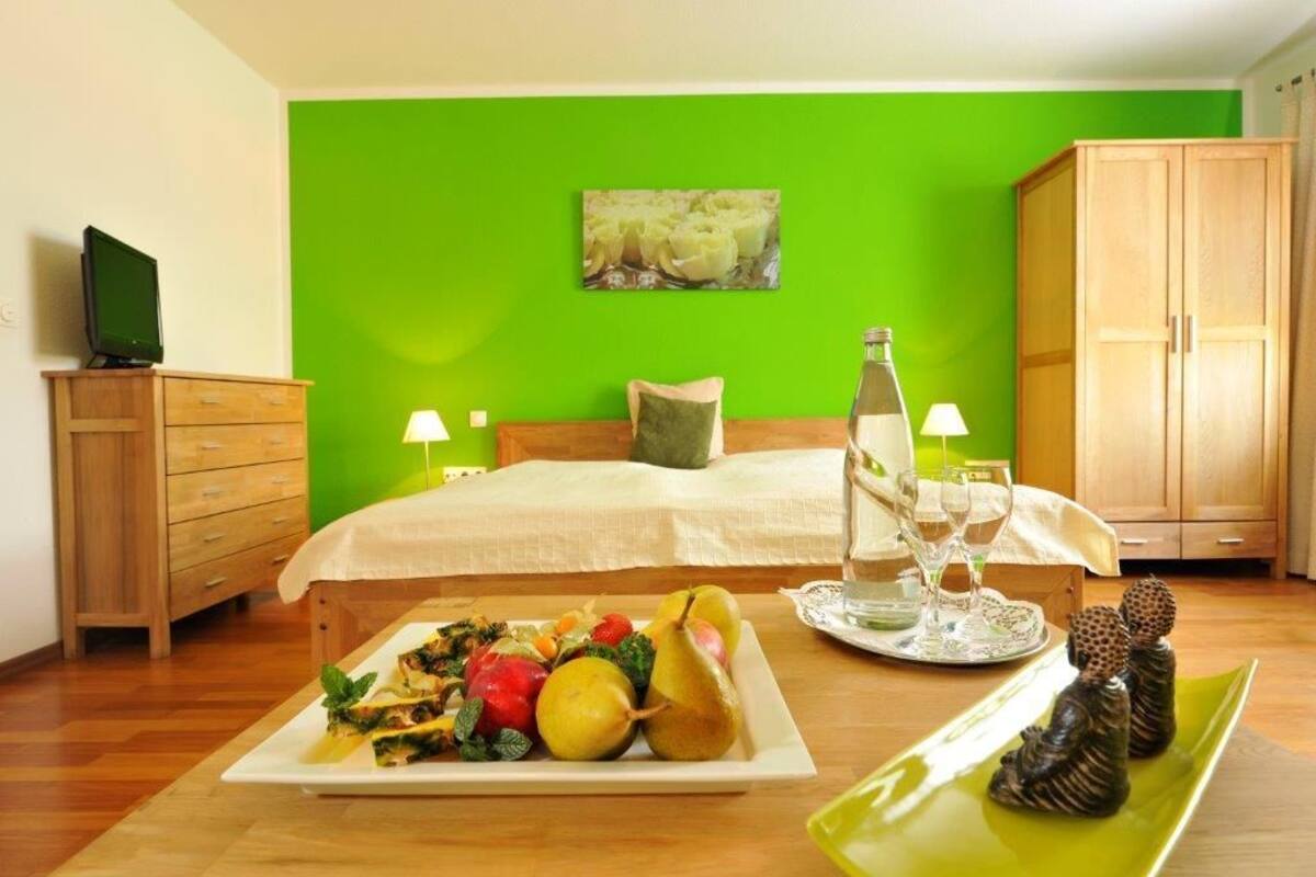 A bedroom with a green wall, a bed with a white bedspread, a wooden dresser, a television on the dresser, a bedside table with a bottle and glasses, and a plate of fruit on the floor.
