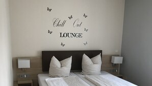 Uma cama de casal com roupa de cama branca, duas mesas de cabeceira com abajures e uma parede com o letreiro 'Chill Out Lounge'.